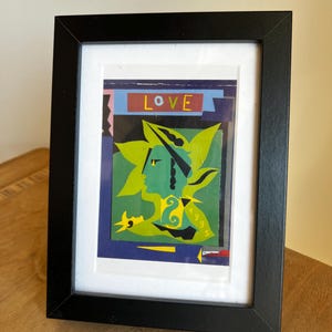 Peut inclure: Impression d'art encadrée avec un visage vert stylisé et des accents jaunes, le mot "LOVE" en rouge. L'œuvre est sur un fond bleu et noir, encadrée dans un cadre noir avec un passe-partout blanc.