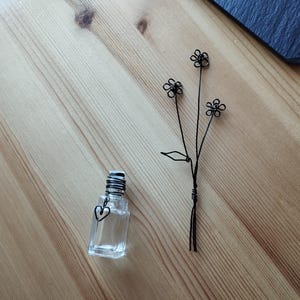May include: A clear glass bottle with a heart-shaped charm and a black wire flower bouquet. The bouquet has three flowers with five petals each.