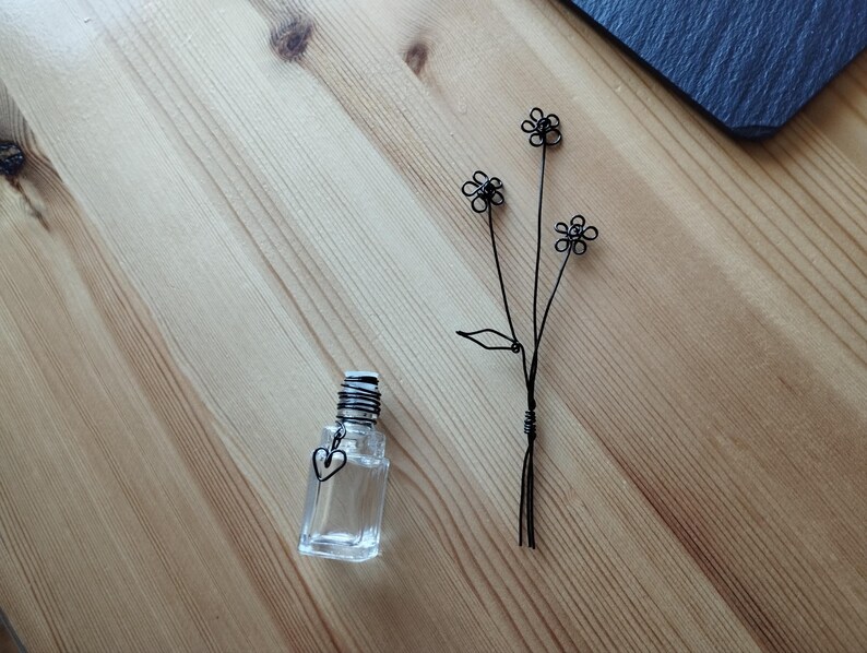 May include: A clear glass bottle with a heart-shaped charm and a black wire flower bouquet. The bouquet has three flowers with five petals each.