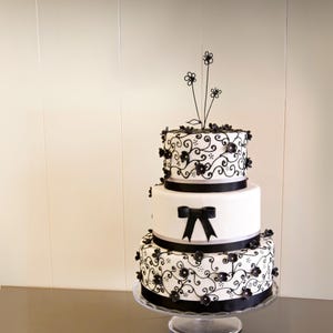 May include: A three-tiered wedding cake decorated with black fondant flowers and intricate scrollwork. The cake is white with black ribbon accents and a black wire flower topper.