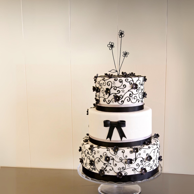 May include: A three-tiered wedding cake decorated with black fondant flowers and intricate scrollwork. The cake is white with black ribbon accents and a black wire flower topper.