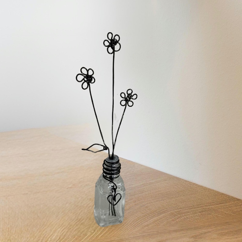 A wire sculpture of three black daisy flowers in a clear glass bottle with a textured surface. The flowers are made of thin wire and are arranged in a vase decorated with a black wire heart.