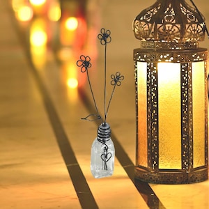 May include: A clear glass vase with a textured surface holds a wire sculpture of black flowers. The vase is sitting on a wooden surface in front of a decorative gold lantern.