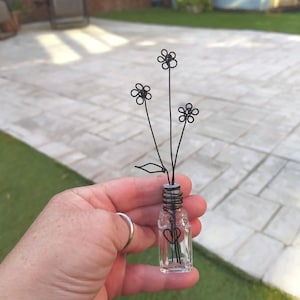 A small glass bottle with a wire-wrapped top with a heart charm, holding a wire sculpture of three black daisy flowers with leaves.