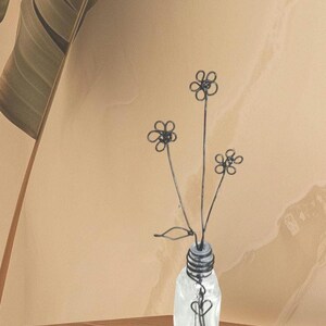 May include: A wire sculpture of flowers in a clear glass vase. The flowers are made of thin wire and are shaped like daisies. The vase is small and has a textured surface.
