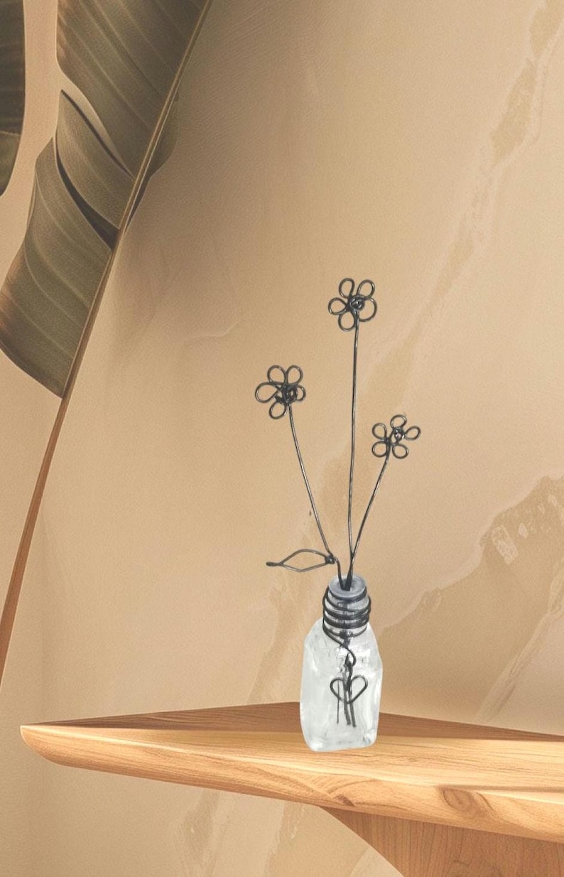 May include: A wire sculpture of flowers in a clear glass vase. The flowers are made of thin wire and are shaped like daisies. The vase is small and has a textured surface.
