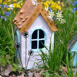A small white cottage with a brown roof and a blue window. The cottage is surrounded by green grass and white flowers. At the front of the house there is a bouquet of three black daisy flowers