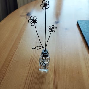 May include: A small glass vase with a black wire flower arrangement. The flowers are simple and delicate, with a heart-shaped design at the base of the vase.