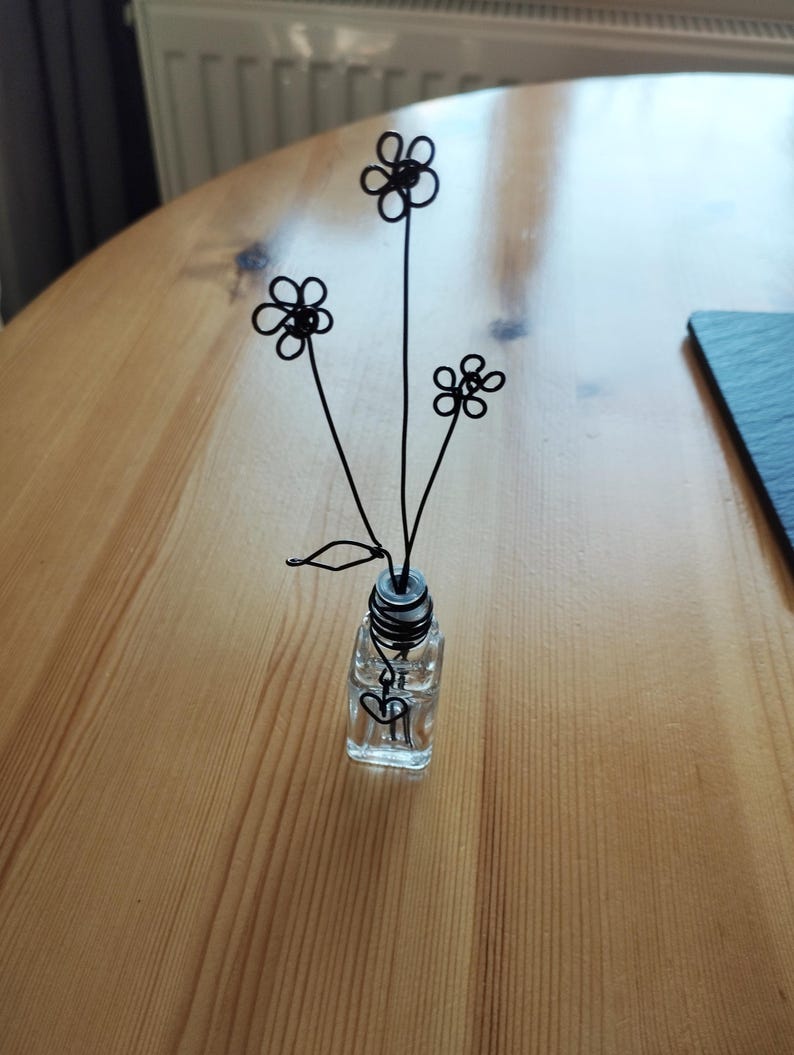 May include: A small glass vase with a black wire flower arrangement. The flowers are simple and delicate, with a heart-shaped design at the base of the vase.