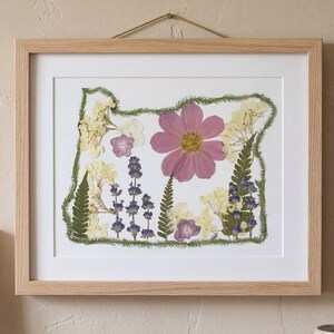 May include: Framed artwork featuring a pressed flower arrangement in the shape of the state of Oregon. The artwork includes a large pink flower, purple and yellow flowers, and green foliage, all set against a white background within a light wood frame.
