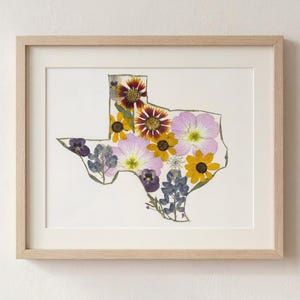 May include: A framed artwork featuring a floral arrangement in the shape of the state of Texas. The artwork showcases a variety of pressed flowers in shades of purple, yellow, and white, set against a white background within a light wood frame.