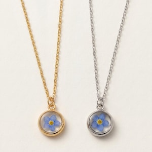 May include: Two necklaces with thin chains, one gold-toned and one silver-toned. Each features a circular pendant containing a single pressed blue forget-me-not flower with a yellow center encased behind a clear, smooth, rounded glass surface.