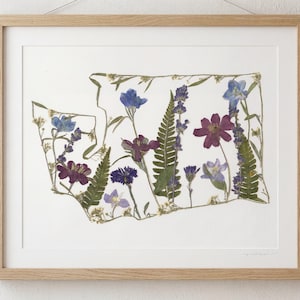 May include: Framed artwork featuring a floral arrangement in the shape of the state of Washington. The artwork includes pressed flowers in shades of purple, blue, and green, set against a white background. The frame is made of light-colored wood.