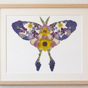 May include: A framed artwork featuring a butterfly design made from pressed flowers. The butterfly's wings are composed of purple, white, and yellow flowers, with a yellow sunflower at the center. The artwork is set against a white background within a light wood frame.