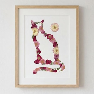 May include: A framed art print featuring a cat silhouette composed of pressed flowers. The flowers are in shades of pink, red, and yellow, set against a white background. The frame is made of light-colored wood.