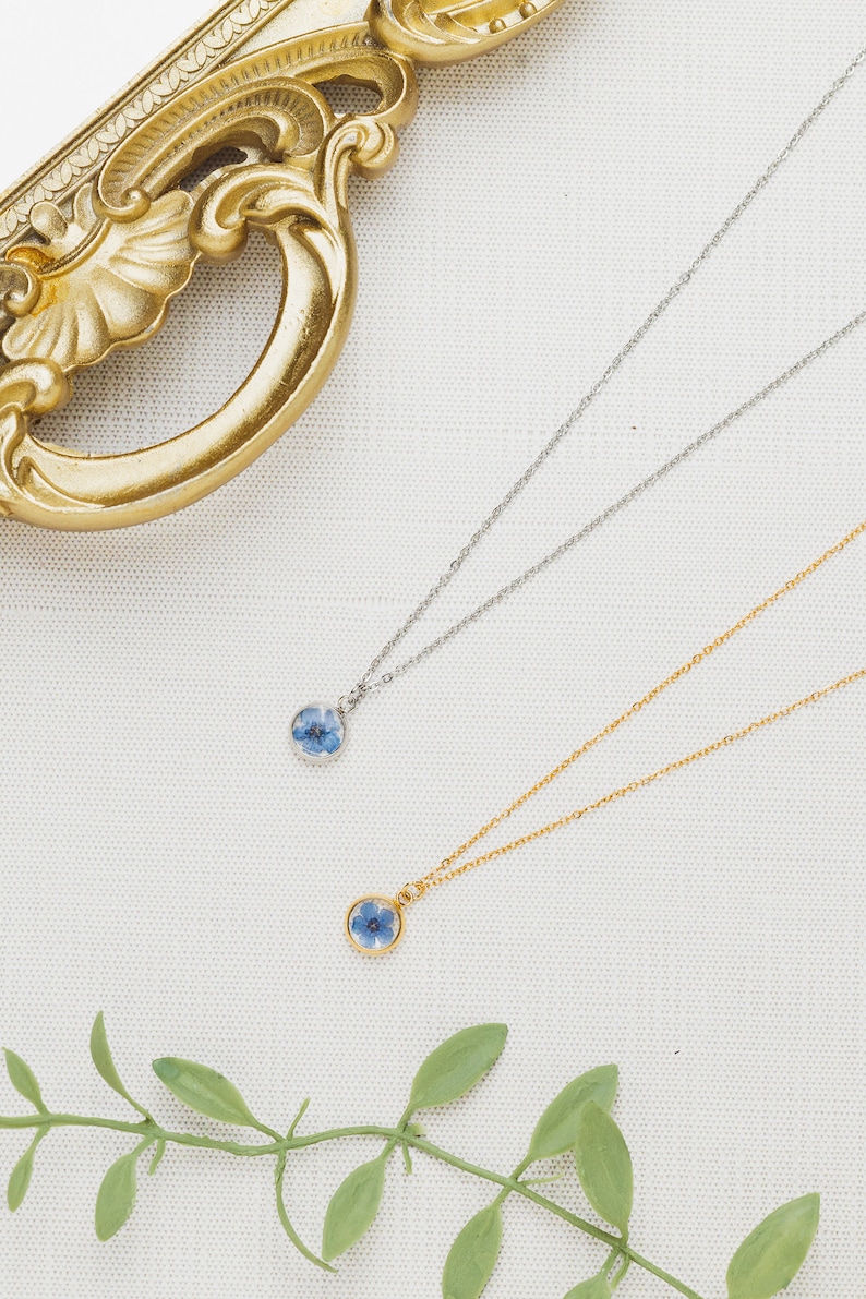 May include: Two delicate necklaces with small, round pendants featuring pressed blue flowers. The necklaces are displayed on a white fabric background with a gold decorative frame and green leaves.