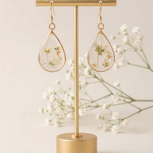 May include: Teardrop-shaped earrings with dried flowers and gold accents. The clear resin earrings are displayed on a gold stand, with a sprig of white flowers in the background. The earrings have gold-colored hooks.