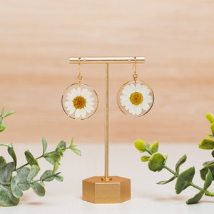 May include: A pair of gold hoop earrings with a white daisy pressed inside each hoop. The earrings are displayed on a gold stand against a light wood background.