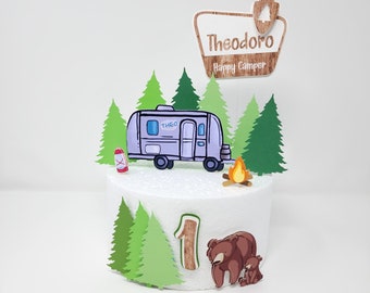 May include: A white cake decorated with a purple camper, green trees, a campfire, a brown bear and a brown bear cub, and a wooden sign that says "Theodoro Happy Camper".