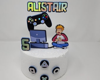 May include: A white cake decorated with a gamer theme. The cake has a blue and black video game controller, a blue computer monitor, a cartoon character playing a video game, and the name Alistair. The cake also has a blue number 5 and blue controller buttons.
