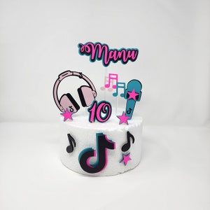 Puede incluir: Un pastel blanco decorado con adornos rosas y azules. El pastel tiene un logotipo de TikTok rosa y negro, un número 10 rosa, un micrófono azul con una estrella rosa, unos auriculares rosas y negros y una nota musical rosa y negra. El pastel también tiene el nombre "Manu" escrito en letras rosas y azules.