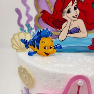 May include: A yellow and blue cartoon fish with a smiling face, a blue fin, and a yellow body. The fish is on a white cake with a purple and gold mermaid figure in the background.