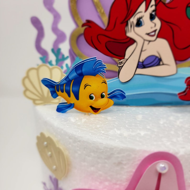 May include: A yellow and blue cartoon fish with a smiling face, a blue fin, and a yellow body. The fish is on a white cake with a purple and gold mermaid figure in the background.