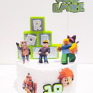 May include: A white cake decorated with a Roblox theme. The cake has green and gray blocks with the letters "R" and "B" on them. There are several Roblox characters on the cake, including a robot, a person with a yellow hard hat, a person with a blue shirt and a cat on their head, and a person with orange hair and headphones. The cake also has a green and gray sign that says "AKIF EMRE" and the number "10" in green and blue.