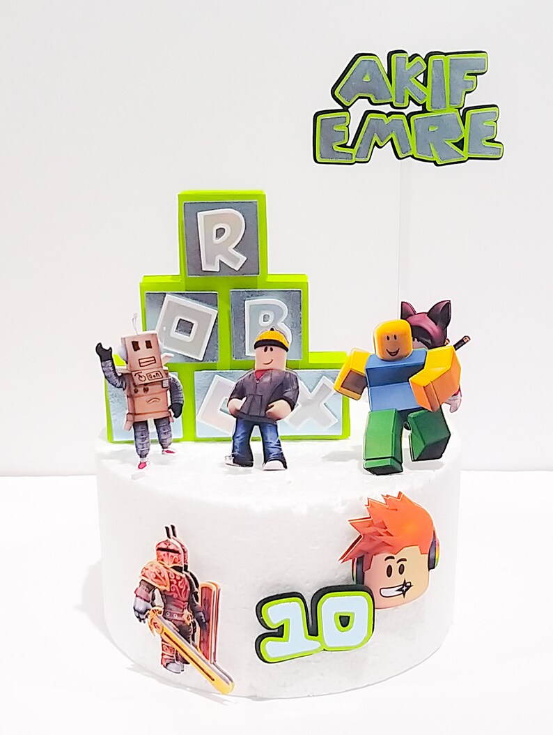 May include: A white cake decorated with a Roblox theme. The cake has green and gray blocks with the letters "R" and "B" on them. There are several Roblox characters on the cake, including a robot, a person with a yellow hard hat, a person with a blue shirt and a cat on their head, and a person with orange hair and headphones. The cake also has a green and gray sign that says "AKIF EMRE" and the number "10" in green and blue.
