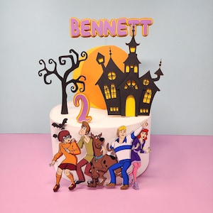 May include: A Scooby Doo themed cake topper with a haunted house, a spooky tree, a bat, and the characters from the show. The topper also includes the name "BENNETT" and the number "2" in purple.