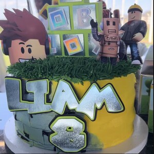 May include: A yellow and gray birthday cake decorated with a green grass border, a silver 'Liam' nameplate, a silver number '8', and Roblox themed decorations including a robot, a figure, and a block with a rainbow pattern.