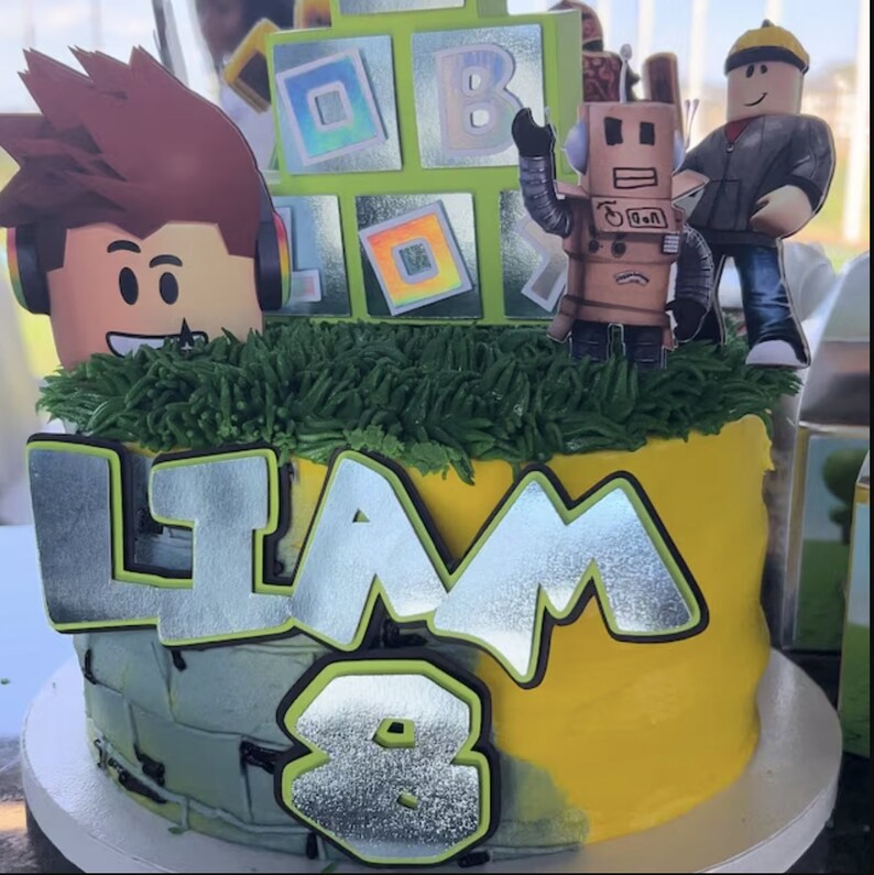 May include: A yellow and gray birthday cake decorated with a green grass border, a silver 'Liam' nameplate, a silver number '8', and Roblox themed decorations including a robot, a figure, and a block with a rainbow pattern.