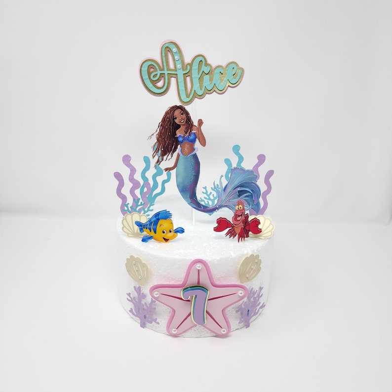 May include: A white cake decorated with a mermaid, a crab, a fish, and a pink starfish with the number 7. The cake also has a topper that says "Alice".