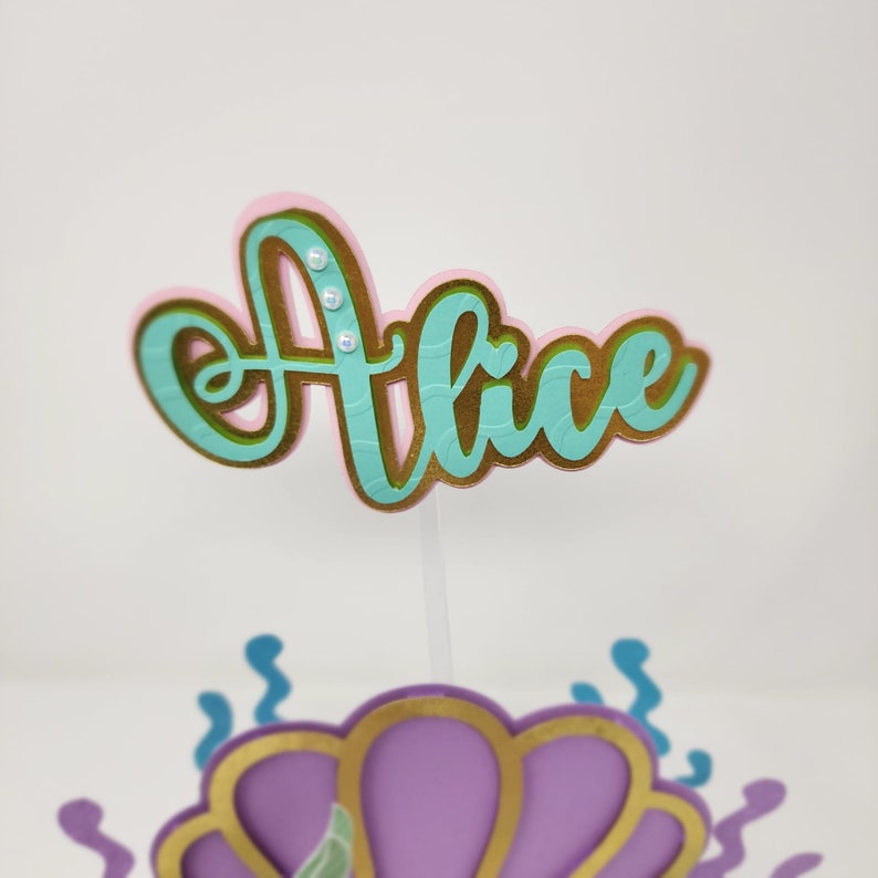 May include: A teal blue and gold cake topper with the name "Alice" in a cursive font. The topper is decorated with small white pearl-like embellishments.