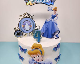 May include: A white cake decorated with a Cinderella theme. The cake has a blue carriage, a Cinderella figurine, a blue butterfly, and the number 4. The cake also has a banner that says "Gabriela".