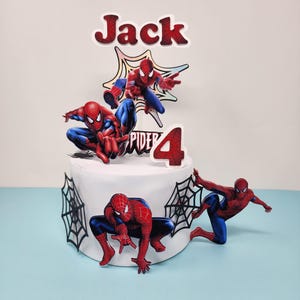 May include: A white cake decorated with Spider-Man themed toppers. The cake features several Spider-Man figures in action poses, a number 4, and the name "Jack". Black spiderweb decorations are also included.