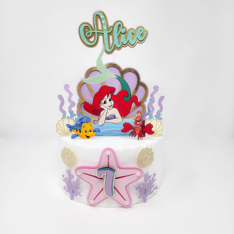 May include: A white cake decorated with a mermaid, a crab, and a fish. The cake is topped with a purple and gold seashell, a green tail, and a gold name topper that says "Alice". The cake also has a pink starfish with a purple number 7 in the center.