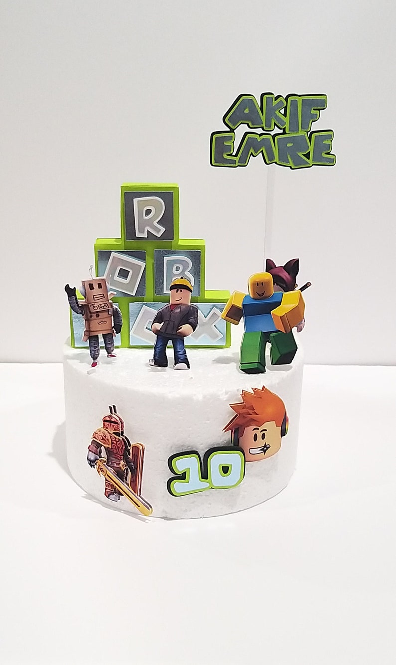 May include: A white cake decorated with Roblox themed figurines, including a robot, a character with a red shirt and a yellow hard hat, a character with a blue shirt and a red hat, and a character with orange hair and headphones. The cake also has a green and white number 10 and a green and white sign that says "AKIF EMRE".