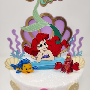 May include: A cake topper featuring Ariel, the Little Mermaid, with her red hair, green tail, and a seashell. The topper also includes a yellow and blue fish and a red crab.