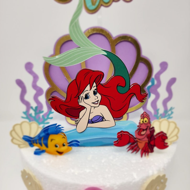 May include: A cake topper featuring Ariel, the Little Mermaid, with her red hair, green tail, and a seashell. The topper also includes a yellow and blue fish and a red crab.
