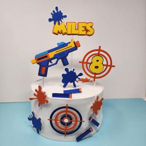 Puede incluir: Una tarta blanca decorada con una pistola de juguete, dianas y salpicaduras de pintura. La decoración de la tarta incluye el nombre "MILES" y el número "8" en una diana. La pistola es azul, amarilla y naranja. Las dianas son rojas, blancas y azules.