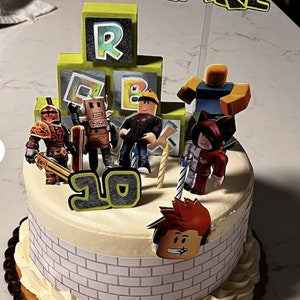 May include: A white cake decorated with a Roblox theme. The cake has green and black Roblox blocks, a red and white figure, a yellow figure, a brown figure, a black and red figure, and a red figure with red hair. The cake also has a number 10 on it.