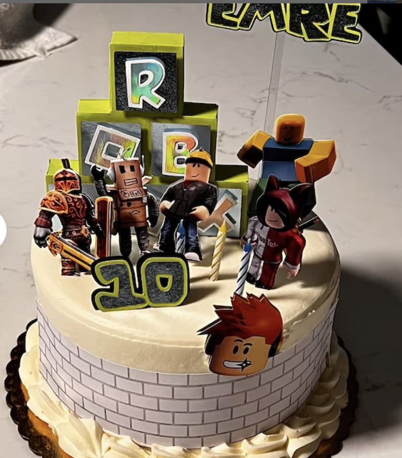 May include: A white cake decorated with a Roblox theme. The cake has green and black Roblox blocks, a red and white figure, a yellow figure, a brown figure, a black and red figure, and a red figure with red hair. The cake also has a number 10 on it.