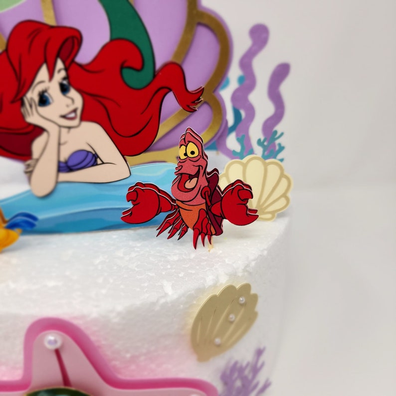 May include: A red and orange cartoon crab with a big smile and open mouth. The crab is standing on a white cake with a purple and gold seashell and a mermaid figure in the background.
