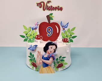 May include: A white cake decorated with a red apple, a number 9, and a Snow White character. The cake also has green leaves, blue birds, and red mushrooms. The cake topper says "Victoria".