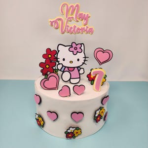 May include: A white cake decorated with pink hearts, flowers, and a Hello Kitty figure. The cake is topped with a pink and yellow sign that reads "May Victoria" and a number 7. The cake is likely for a birthday celebration.