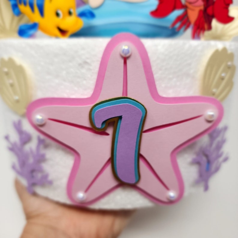 May include: A pink starfish with a purple number 7 in the center. The starfish is decorated with gold and blue accents and has white pearl-like embellishments around the edges.