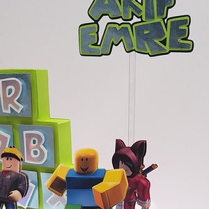 May include: A birthday cake decorated with a Roblox theme. The cake is decorated with green and blue blocks, a Roblox character, and a topper that says "AKIF EMRE".