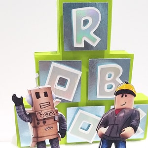 May include: A green and blue cardboard cake topper with the word "ROBLOX" spelled out in white letters. There are two figures on the cake topper, a cardboard robot and a Roblox character.
