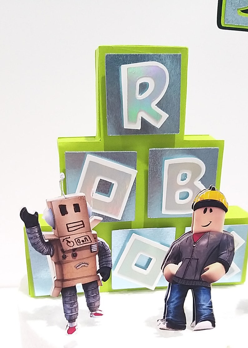 May include: A green and blue cardboard cake topper with the word "ROBLOX" spelled out in white letters. There are two figures on the cake topper, a cardboard robot and a Roblox character.